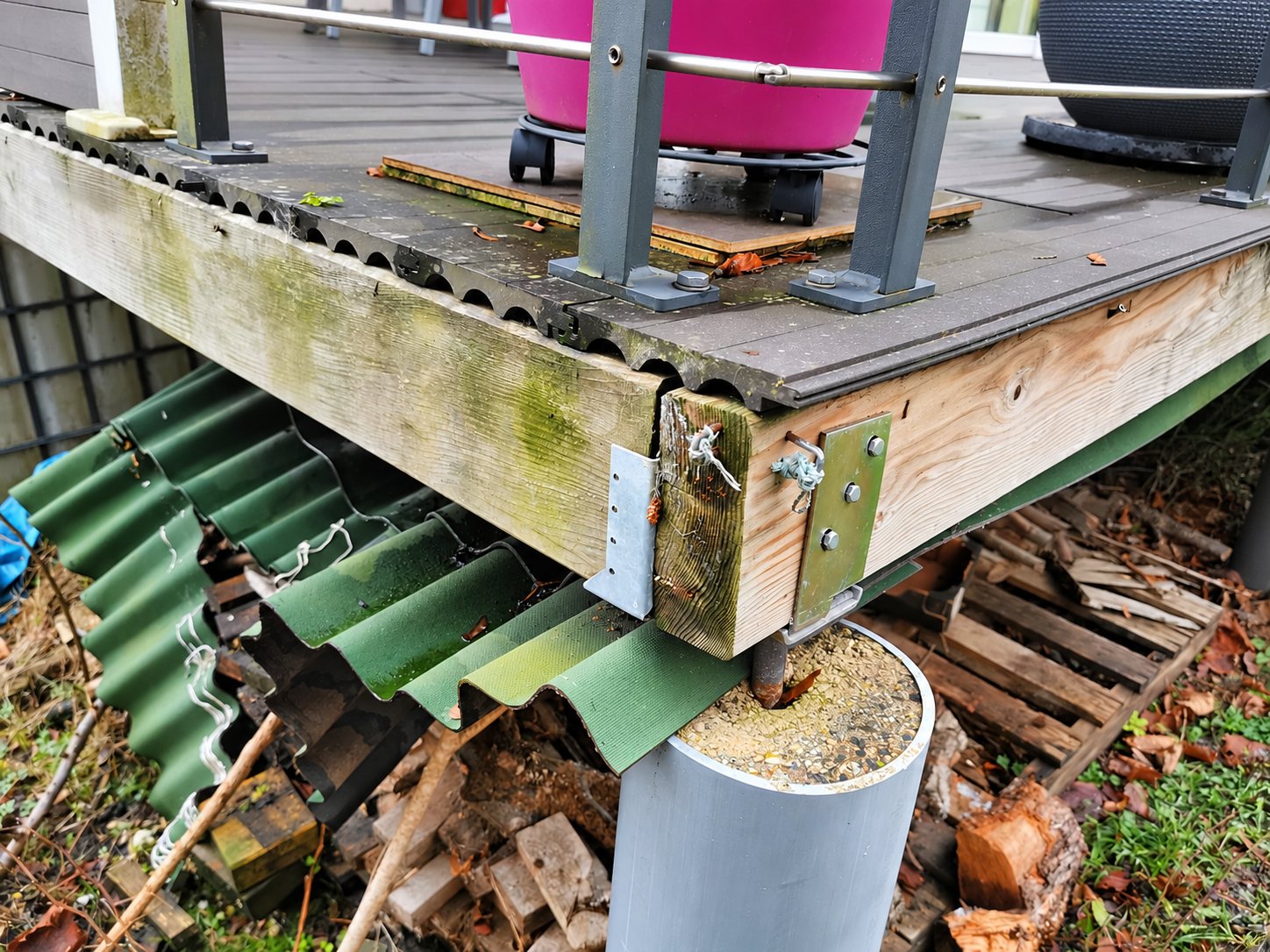 Gros plan sur l'angle d'une terrasse en bois et composite, fixé par des équerres métalliques sur un plot en tube gris, avec des plaques ondulées vertes et du bois empilé en dessous.