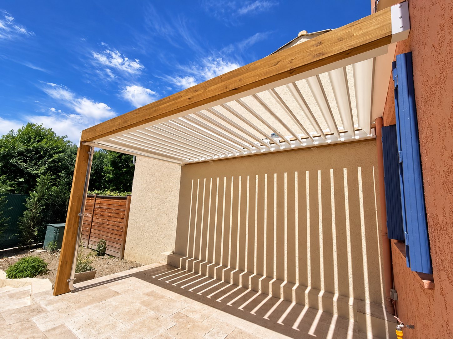 Une pergola en bois à lames orientables blanches projette des rayures de lumière sur le mur beige d'une terrasse dallée, sous un ciel bleu.