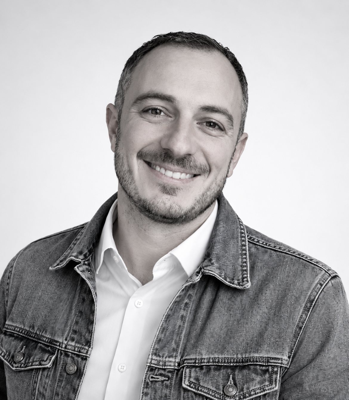 Portrait en noir et blanc d'un homme souriant, barbe courte, portant une chemise blanche sous une veste en jean, devant un fond uni.