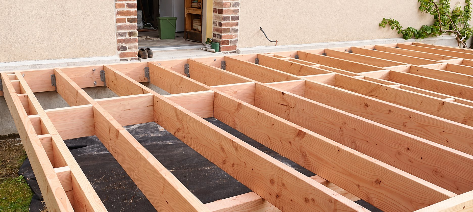 Ossature de terrasse en bois composée de solives en douglas posées sur une bâche noire, devant une façade de maison avec une porte ouverte.